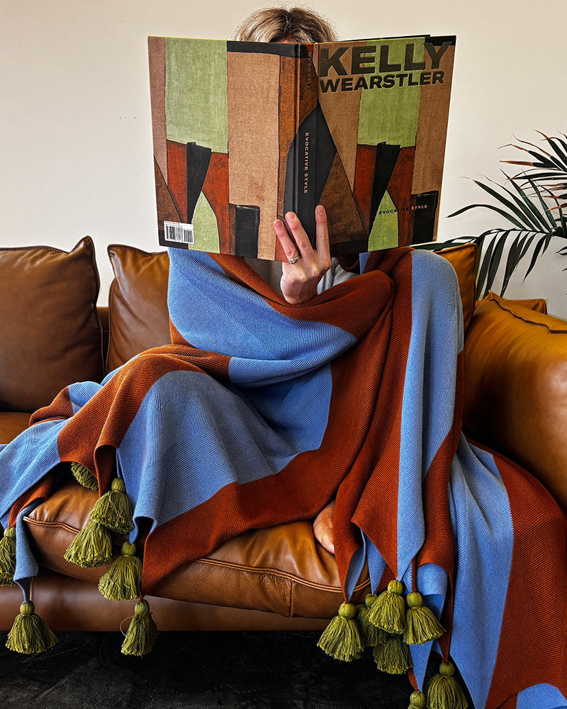 Person sitting on a brown leather couch with a colorful blue and brown stripe blanket with chunky tassels, holding a book titled 'Kelly Wearstler'.