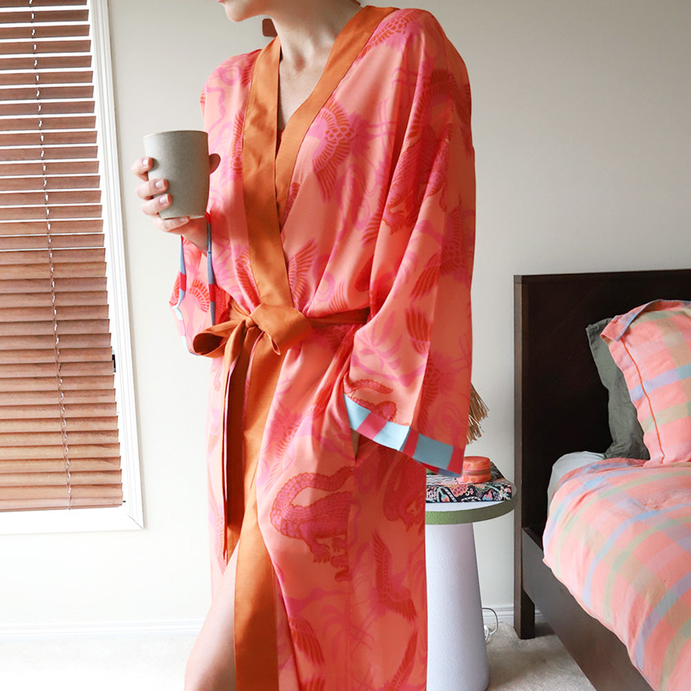 Person wearing a pink and orange summer robe holding a mug in a bedroom setting.