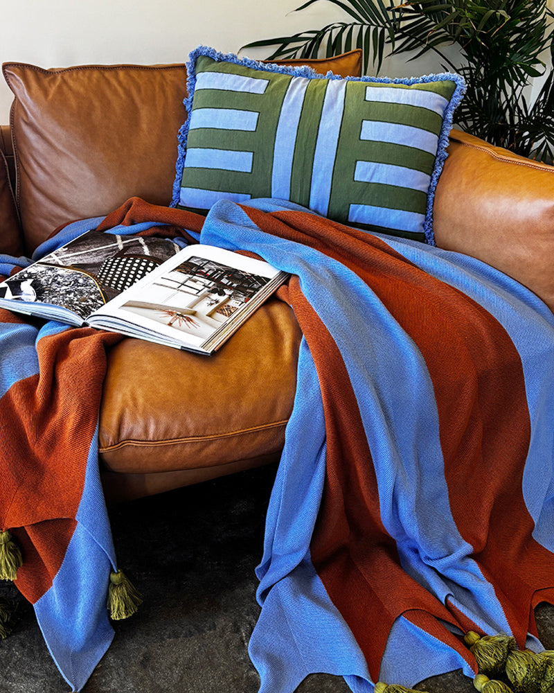 Brown leather sofa with a colorful blanket in brown and blue with chunky olive tassels, magazine, and striped pillow.