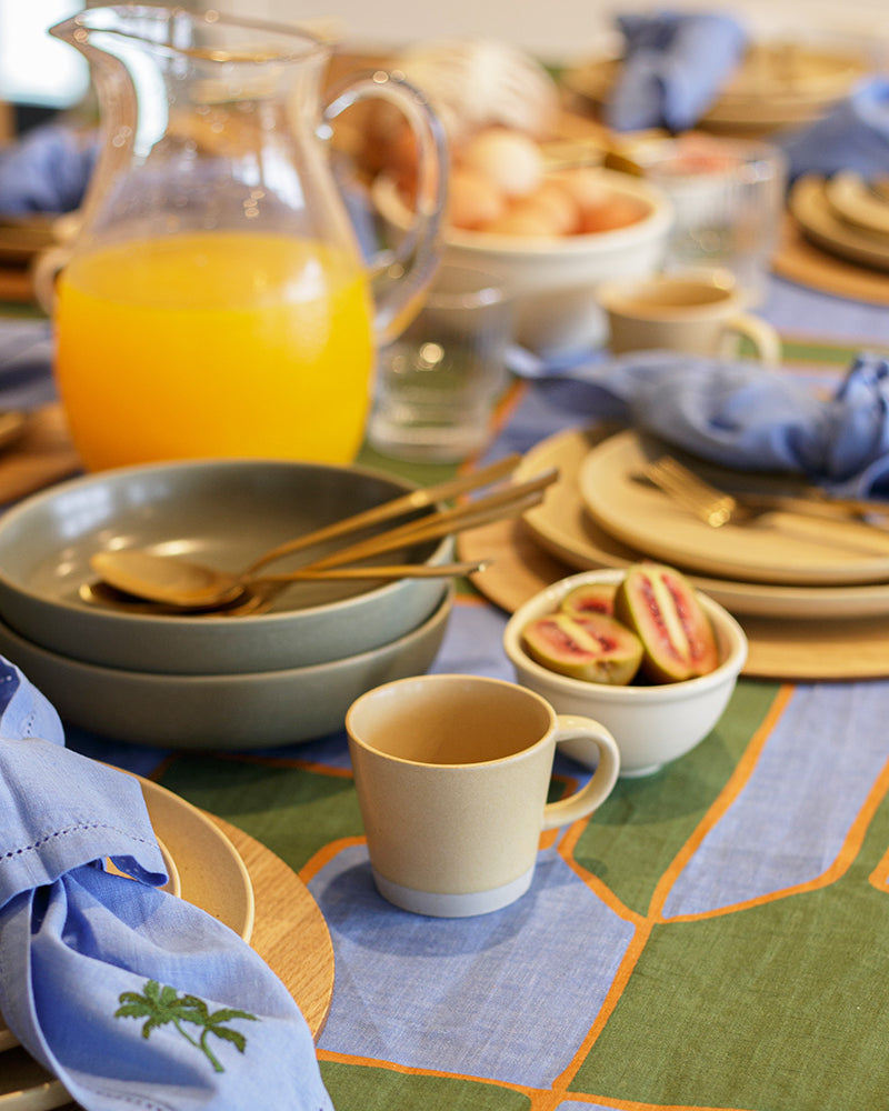 Green and blue dining table set with plates, cups, and a pitcher of orange juice on a patterned linen tablecloth.