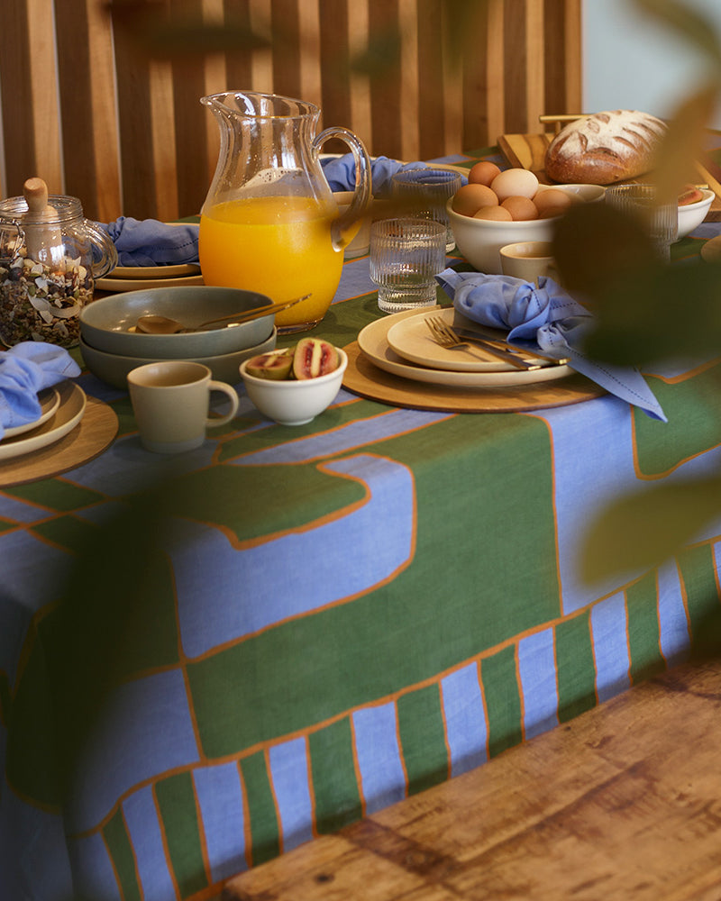 Green and blue dining table setting for slow mornings with plates, cups, and a pitcher of orange juice on a patterned linen tablecloth.