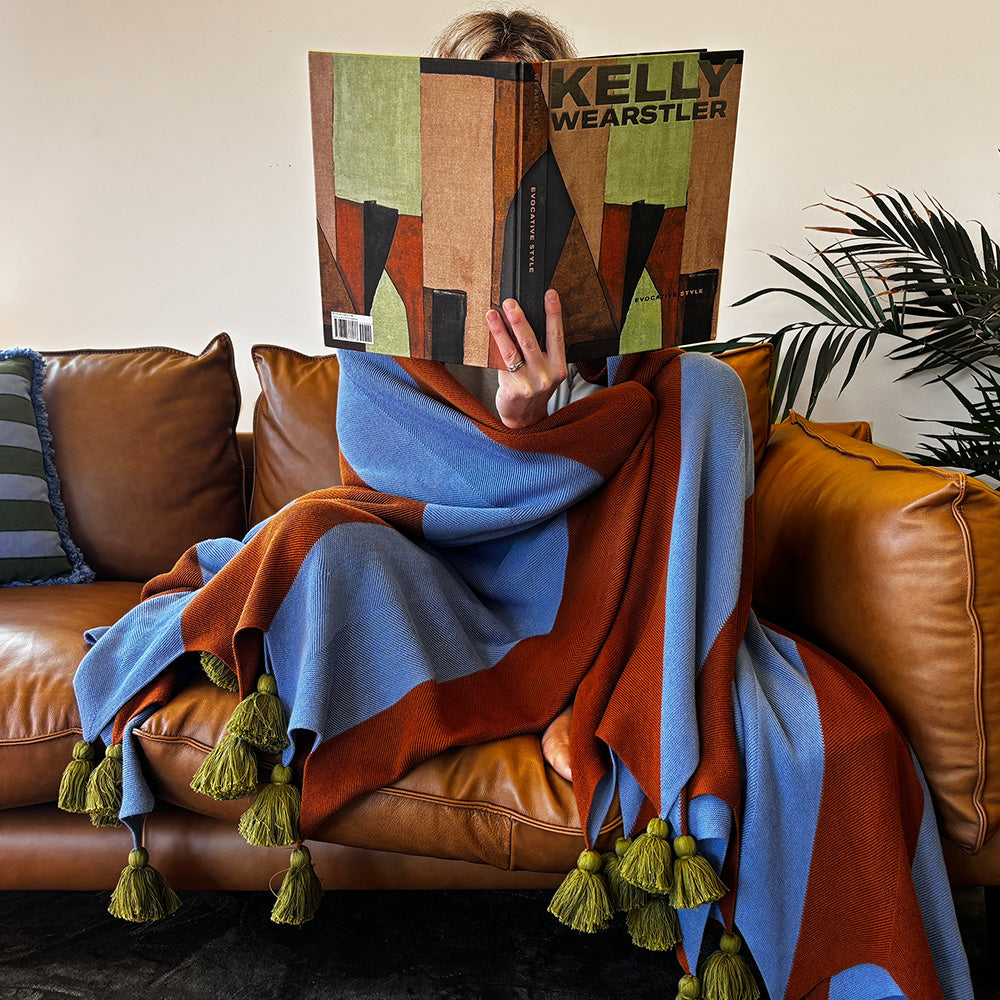 Person sitting on a brown leather couch with a colorful  blue and brown stripe blanket with chunky tassels, holding a book titled 'Kelly Wearstler'.