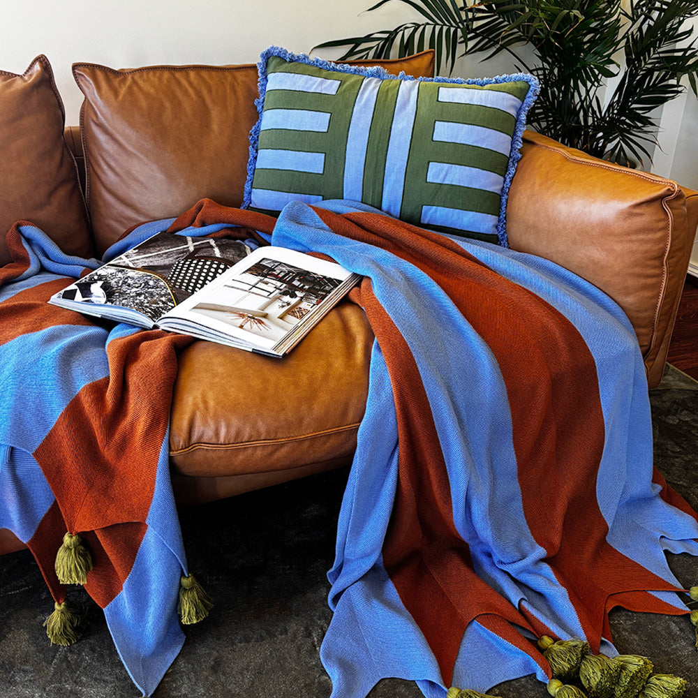 Brown leather sofa with a colorful blanket in brown and blue with chunky olive tassels, magazine, and striped pillow.