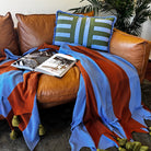 Brown leather sofa with a colorful blanket in brown and blue with chunky olive tassels, magazine, and striped pillow.