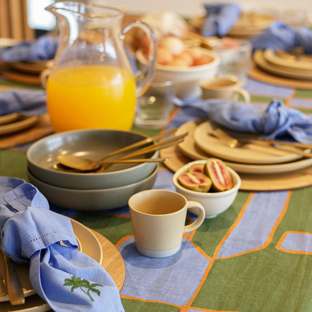 Green and blue dining table set with plates, cups, and a pitcher of orange juice on a patterned linen tablecloth.
