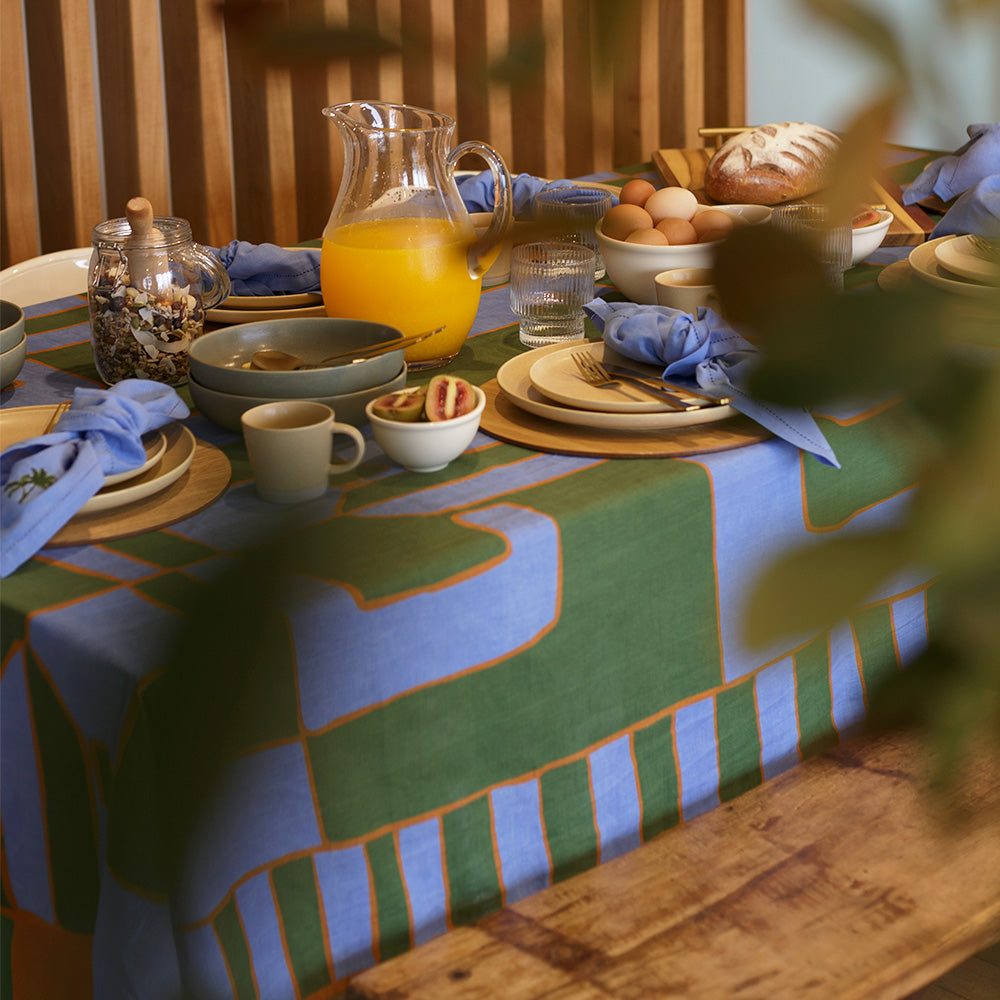 Green and blue dining table setting for slow mornings with plates, cups, and a pitcher of orange juice on a patterned linen tablecloth.