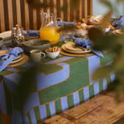 Green and blue dining table setting for slow mornings with plates, cups, and a pitcher of orange juice on a patterned linen tablecloth.